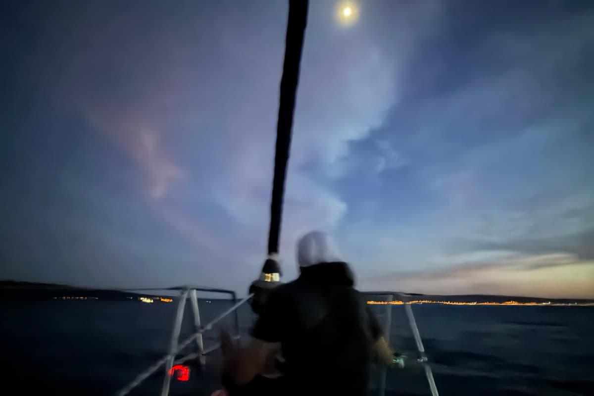 Ein Mann sitzt am Bug des Segelboots unter dem Vollmond, am Horizont die Lichter der Küste