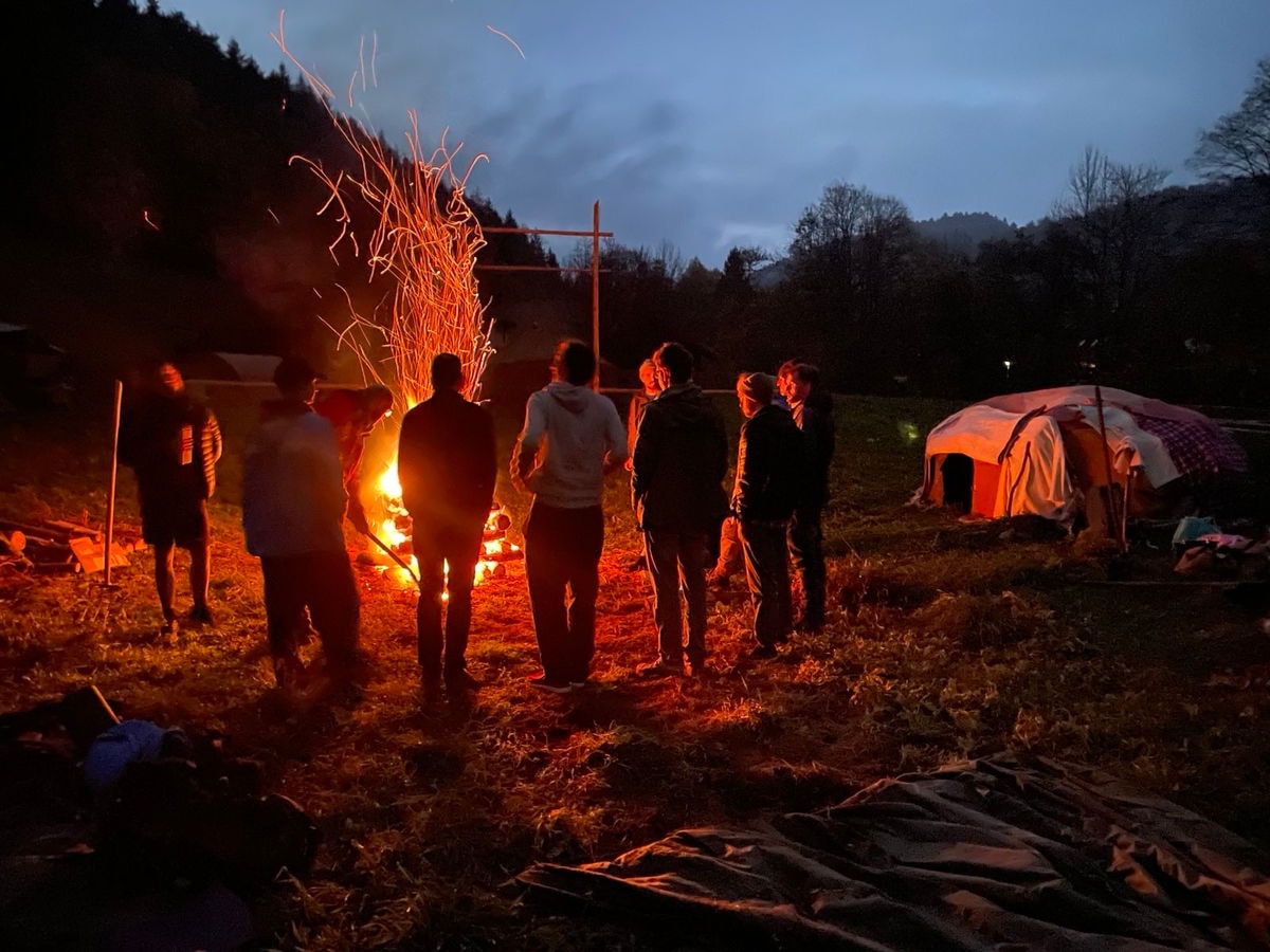 Männer stehen bei Nacht um ein loderndes Feuer, im Hintergrund eine Hütte