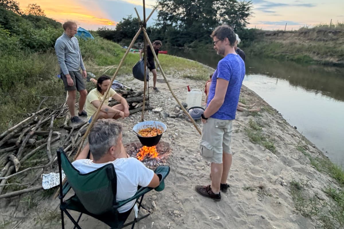 Männer kochen an einem Dreibein über dem Feuer auf einer Sandbank